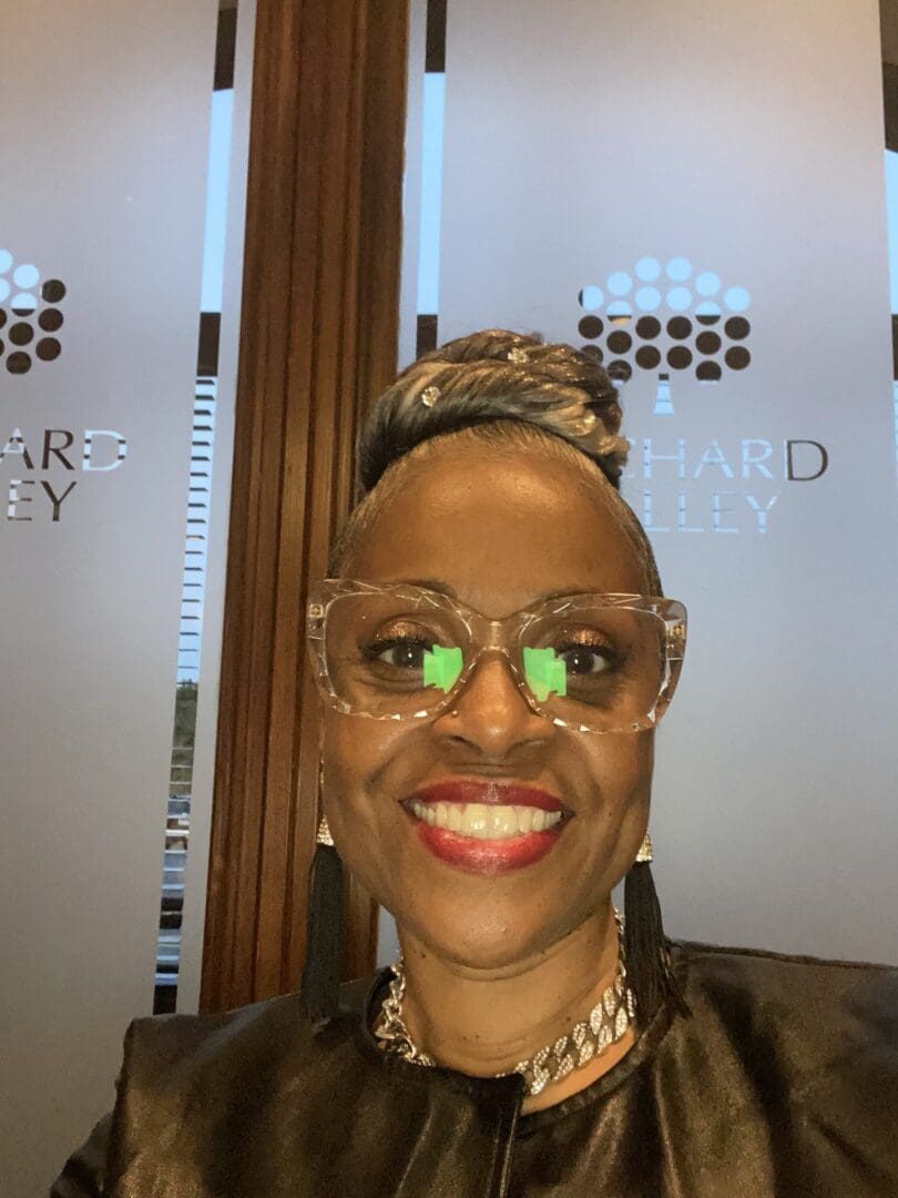 Smiling woman wearing glasses with a wooden background and a logo behind her.